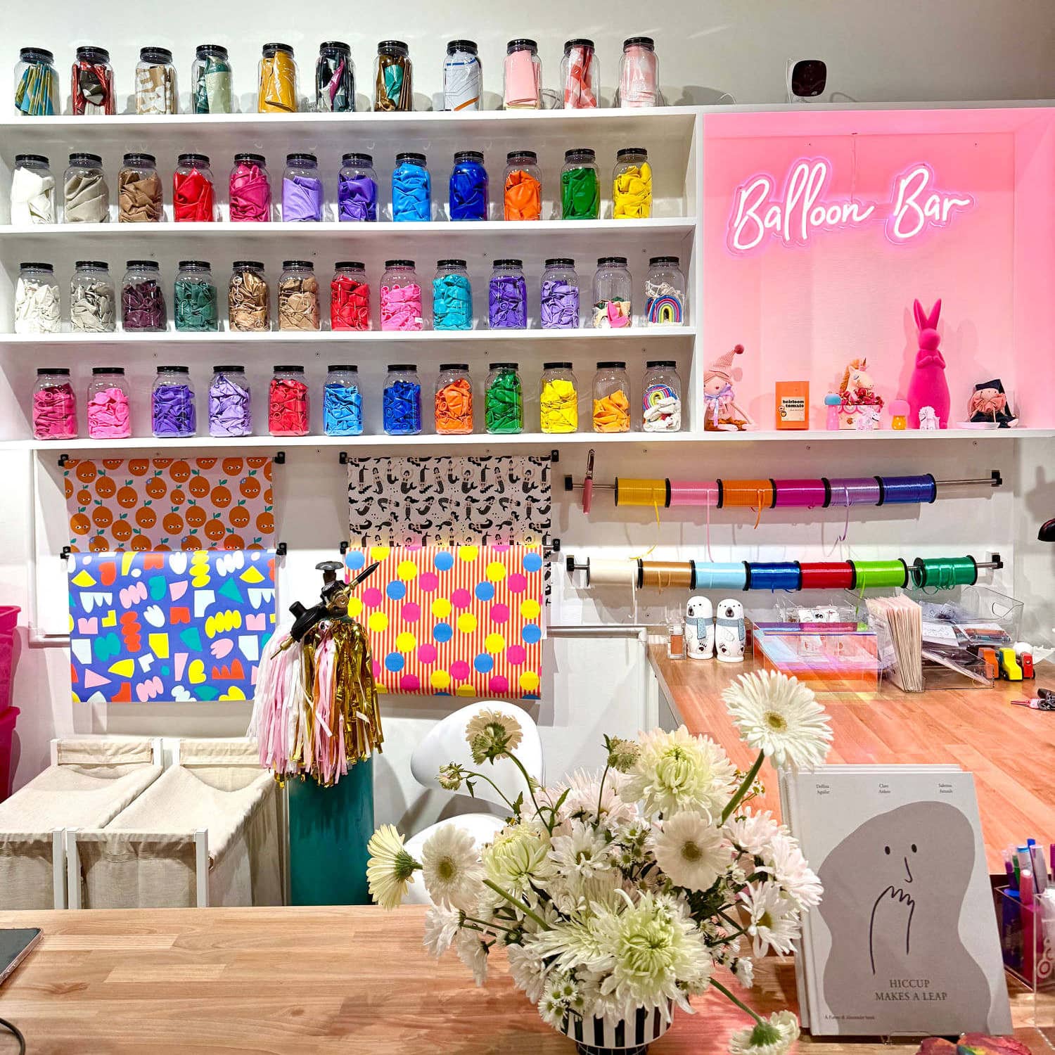The balloon bar: shelves of glass jars holding loose latex balloons sorted by color, ribbon spools on the rail below, and a pink neon Balloon Bar sign glowing beside them