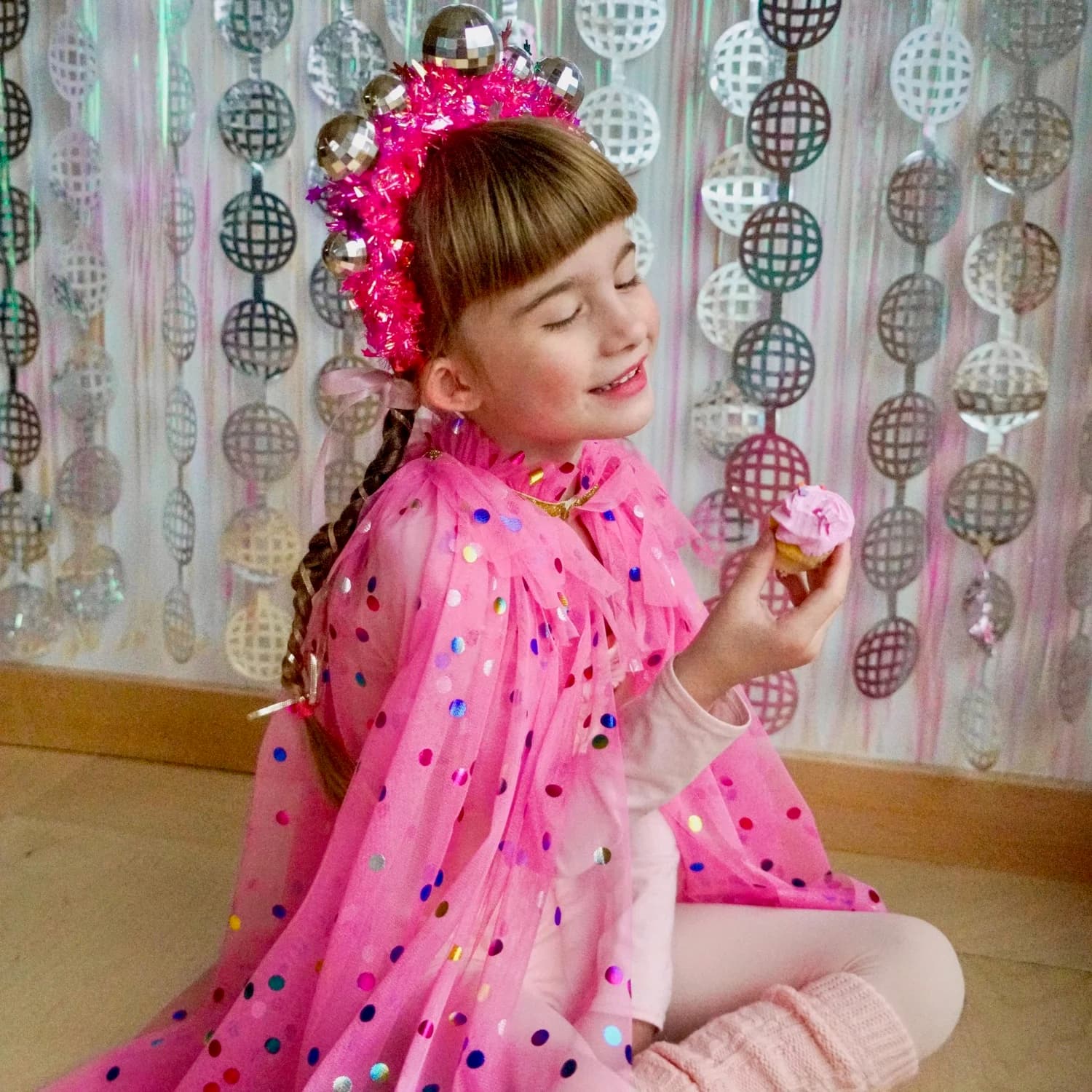 A smiling girl in a pink polka-dot cape and a disco-ball crown holding a frosted cupcake in front of a shimmering disco-ball backdrop