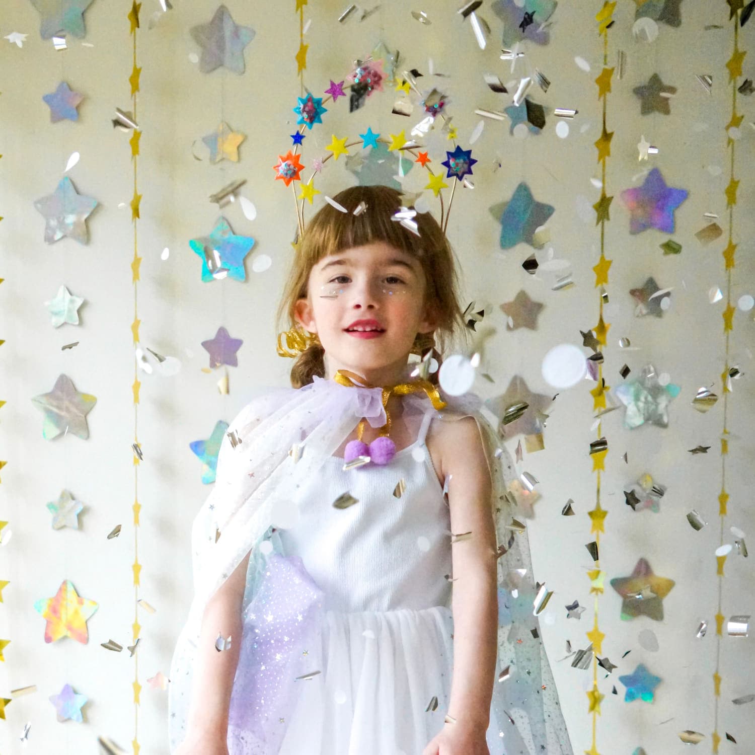 A smiling girl in a halo crown of glitter stars as silver confetti falls around her