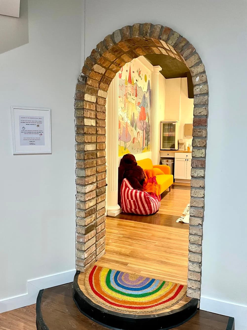 View through the brick archway into the playroom: a yellow couch under a rainbow mural