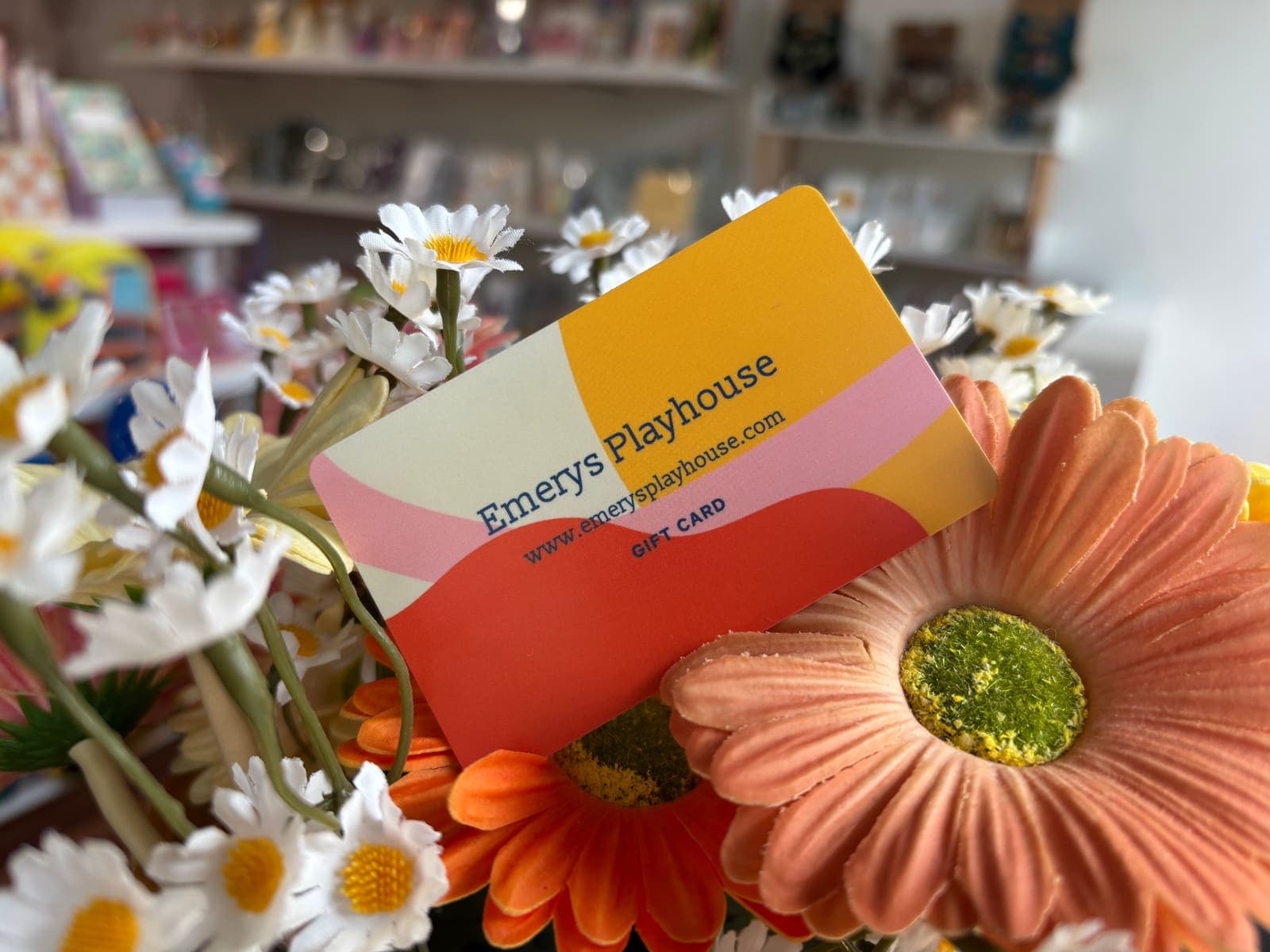 An Emery's Playhouse gift card tucked into a bunch of daisies and gerberas, with the shop's shelves behind it